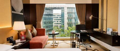 Habitación de hotel con cama, escritorio, silla, lámpara y vistas a edificios a través del ventanal.