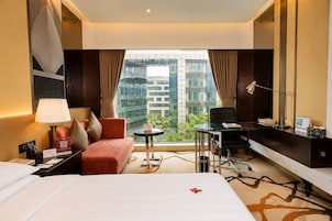 A hotel room with a bed, a desk, a chair, a lamp, and a view of buildings through the window.