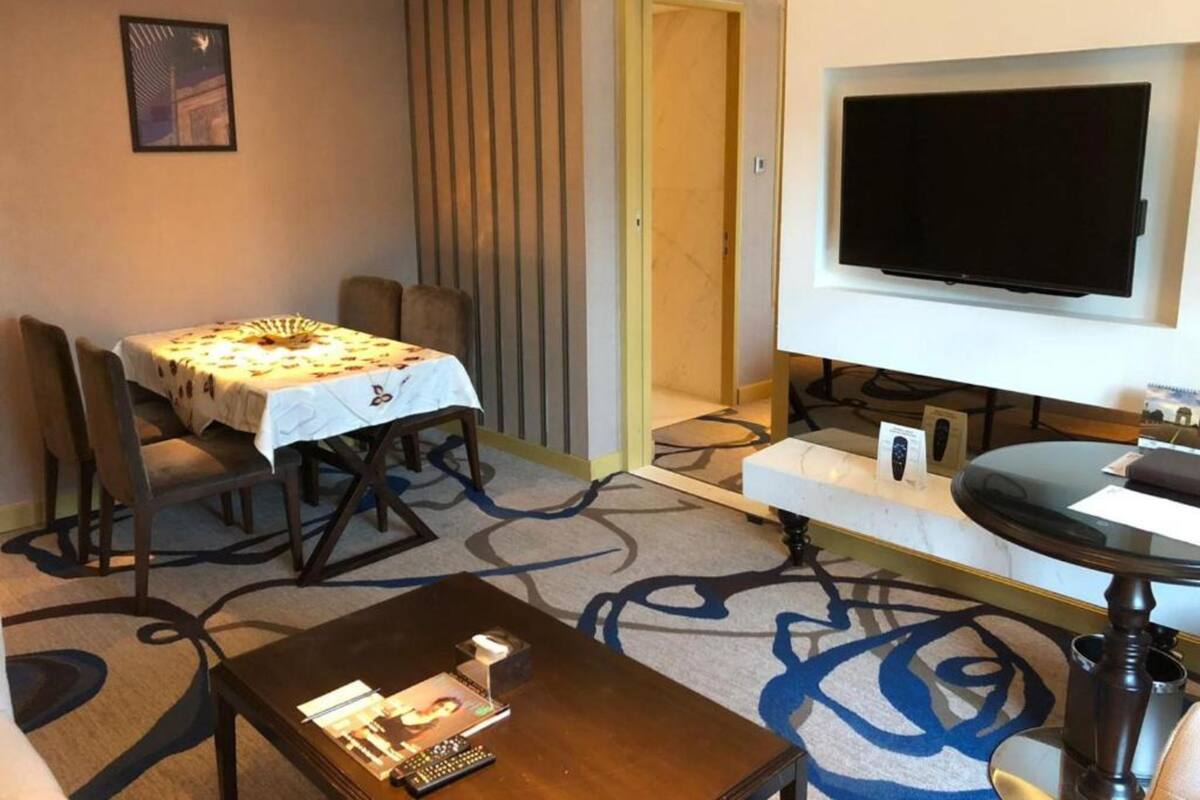 A hotel room with a dining table, chairs, a TV, a coffee table, and a carpet with a blue abstract design.