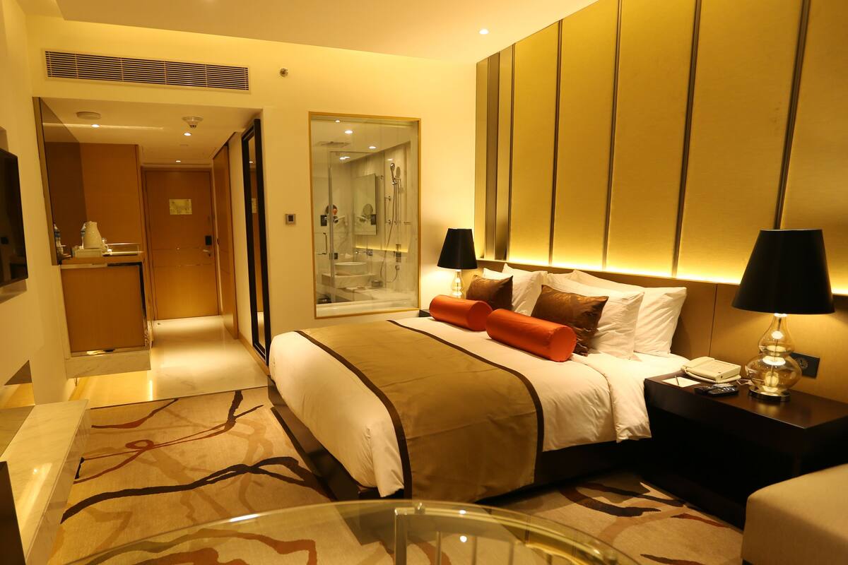 A hotel room with a bed, bedside lamps, a desk, and a bathroom visible through a glass door.