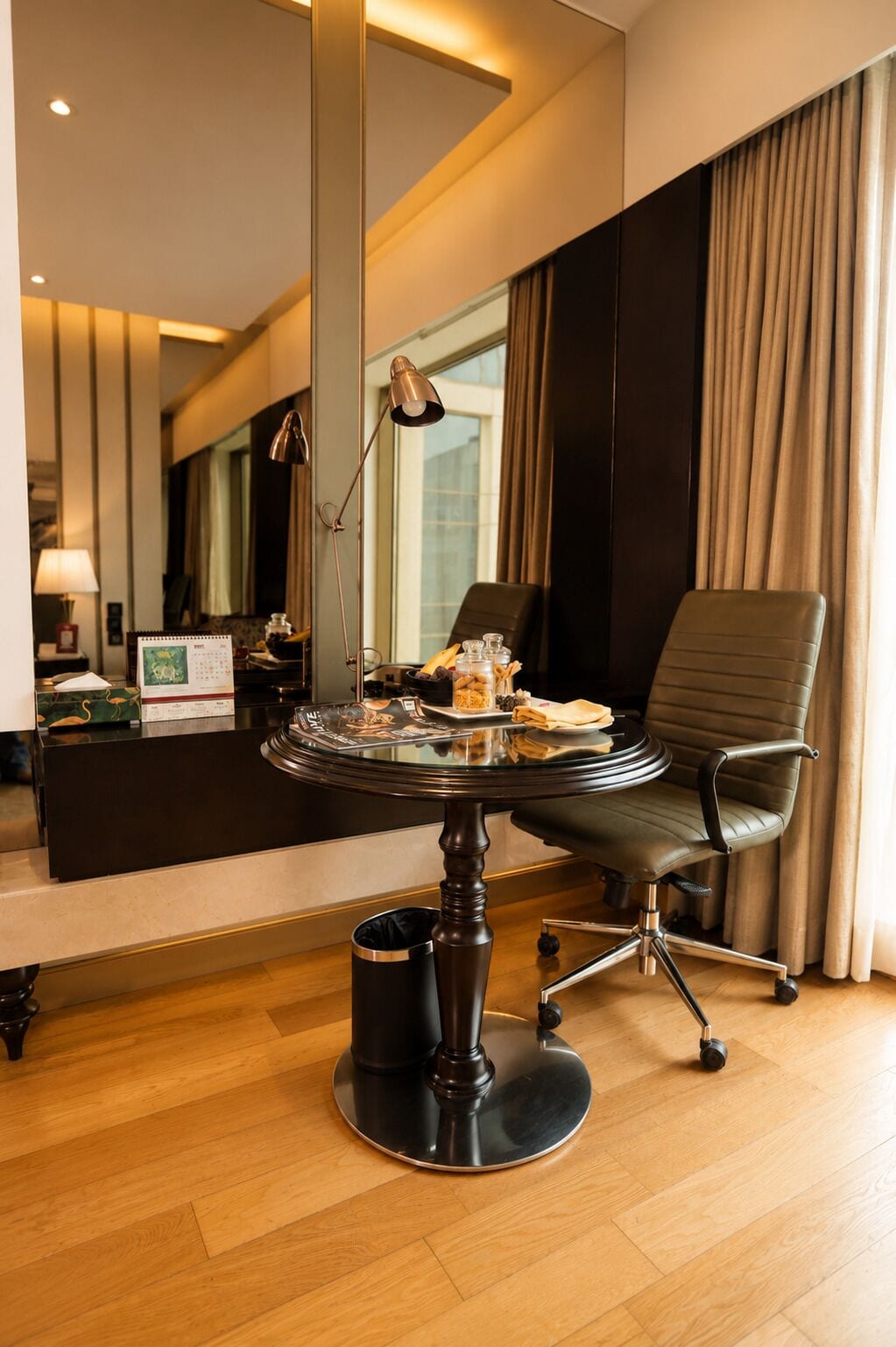 A hotel room with a round table, a leather chair, a desk with a lamp, and a large mirror.