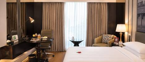 Habitación de hotel con una cama grande, un escritorio con lámpara, una silla, una mesita con lámpara y vistas al exterior a través de la ventana.