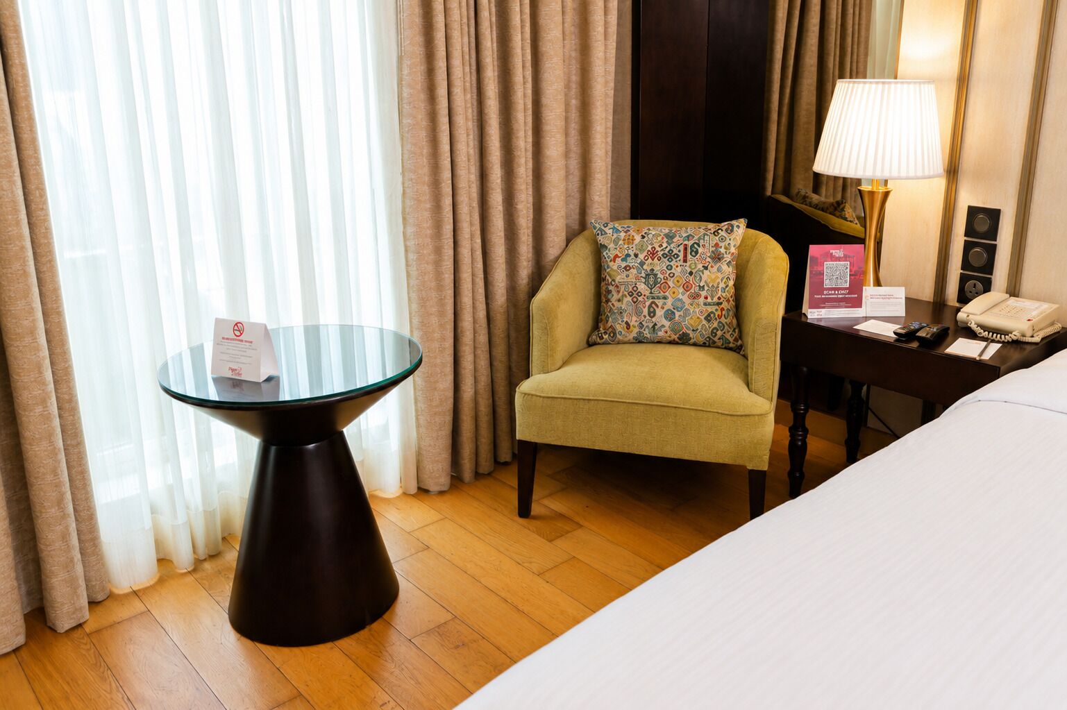 A hotel room with a bed, a round glass table, a chair with a patterned cushion, a lamp, a telephone, and a sign indicating a smoking area.