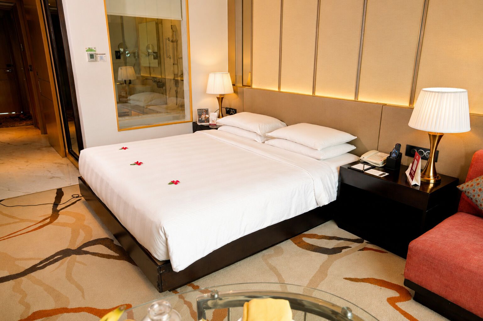 A hotel room with a large bed, two bedside tables, a nightstand, a glass coffee table, and a red armchair.