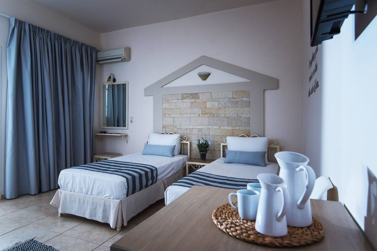 A hotel room with two beds, a wooden table with white pitchers, a stone accent wall, and a window with curtains.
