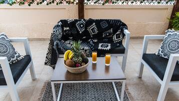 Un balcón con una cesta de mimbre llena de fruta, dos vasos de jugo de naranja y un plaid estampado sobre un sofá.