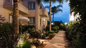 A well-lit pathway leading to a building with a balcony, surrounded by lush greenery and palm trees. - Le Palme (Hersonissos)