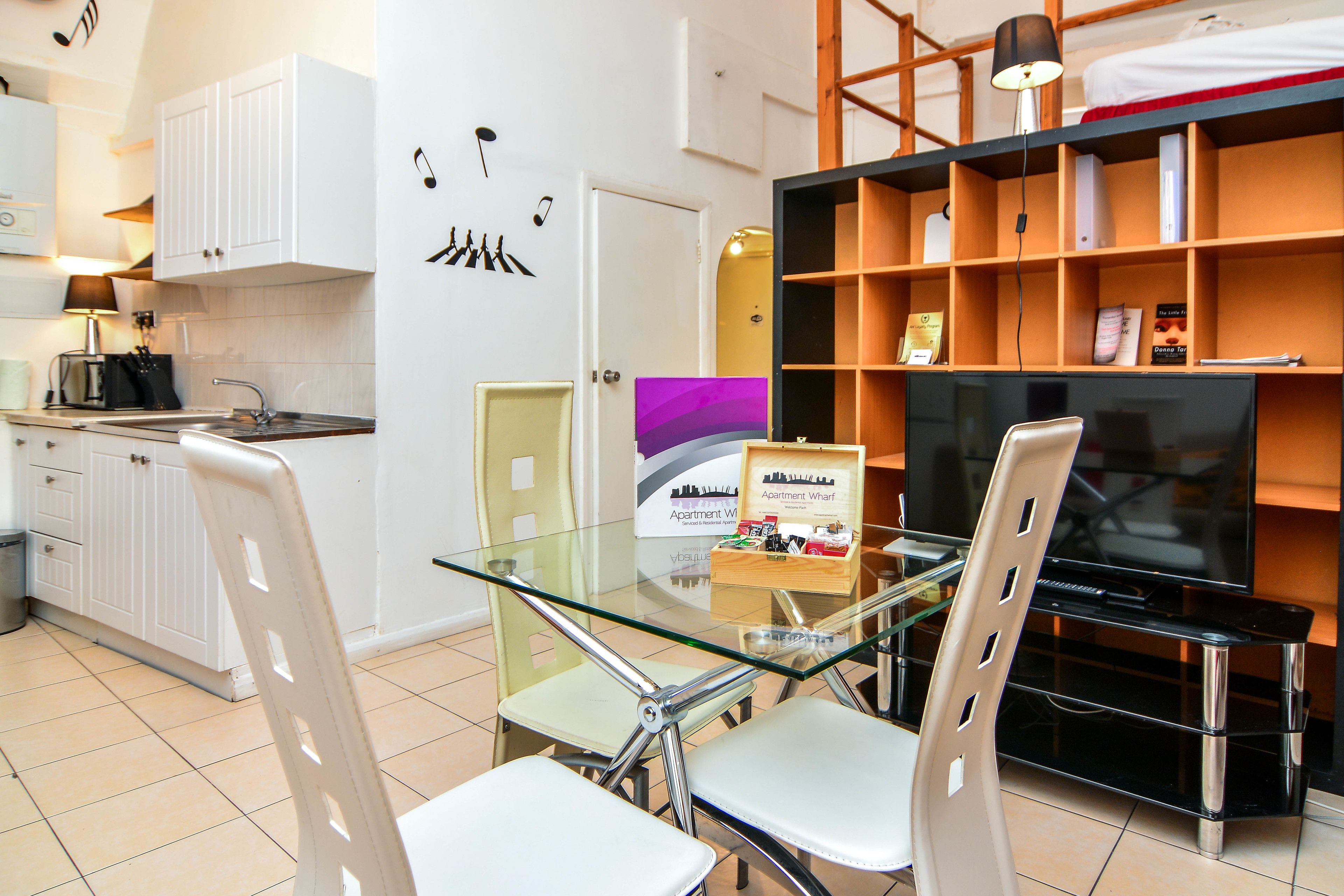 Un espace de vie compact comprenant un coin cuisine, un coin repas avec une table en verre et un téléviseur posé sur une étagère en bois.