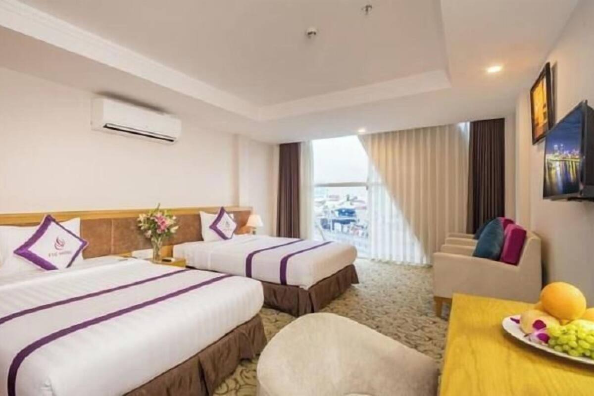 A hotel room with two beds, a sofa, a dining table with fruit and wine, and a TV on the wall.