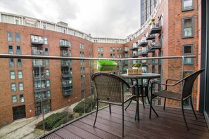 A balcony with wicker chairs and a table, overlooking a brick building with multiple balconies.