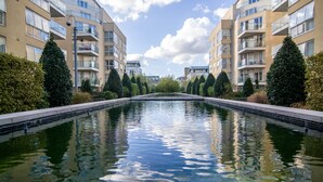 A swimming pool flanked by apartment buildings with balconies, surrounded by neatly trimmed hedges.