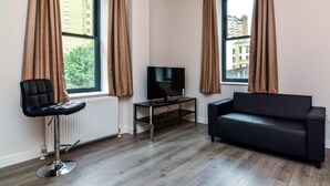 Un salon moderne avec un canapé en cuir noir, un fauteuil en cuir noir, un téléviseur posé sur une table en verre et de grandes fenêtres ornées de rideaux.