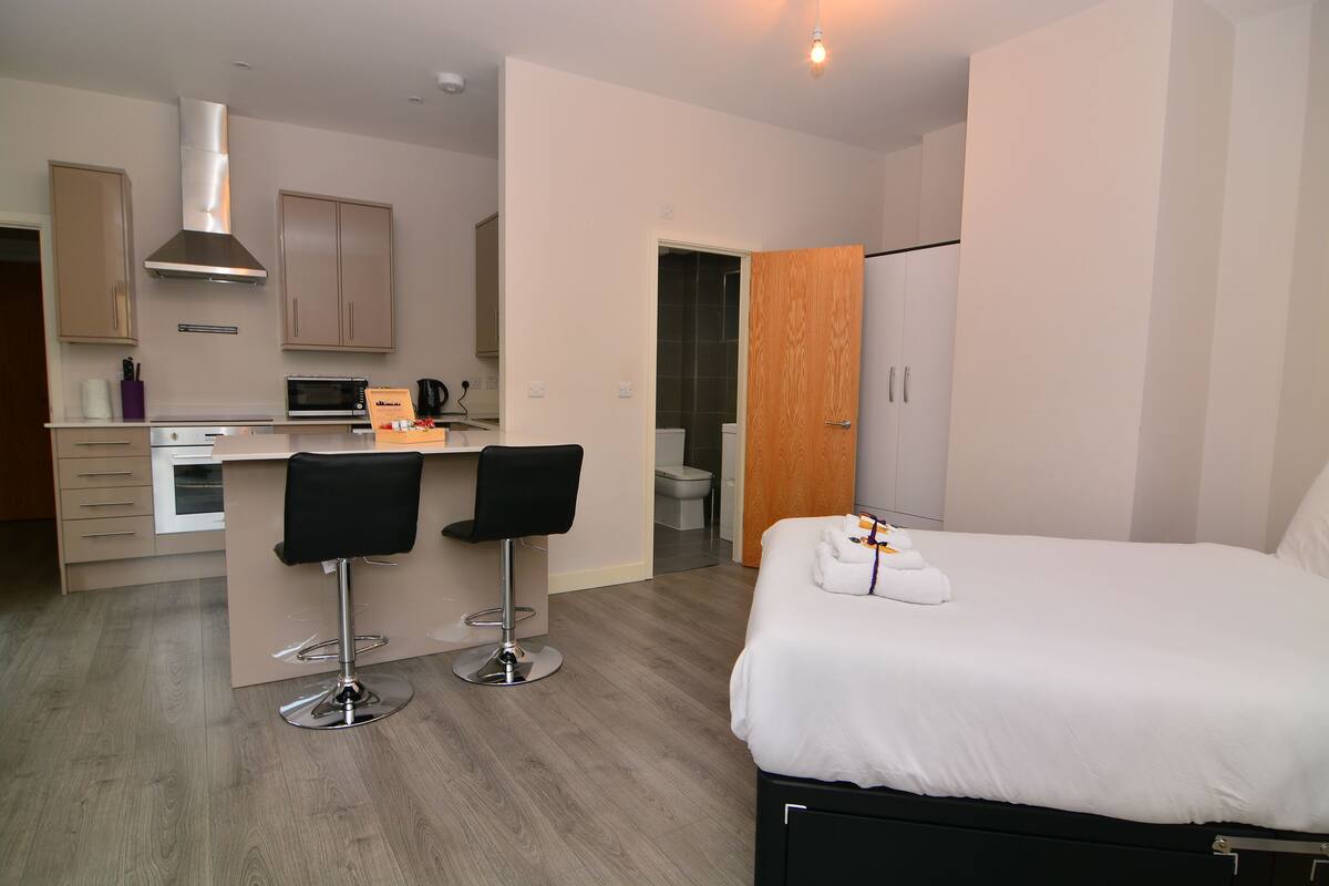 Ein modernes Hotelzimmer mit einem Bett, einer kleinen Küchenzeile, zwei Barhocker und einem Holzschrank.