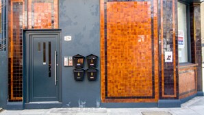 Un immeuble avec une porte de couleur gris foncé, portant le numéro 24, et un mur recouvert de carreaux aux motifs orange et marron.