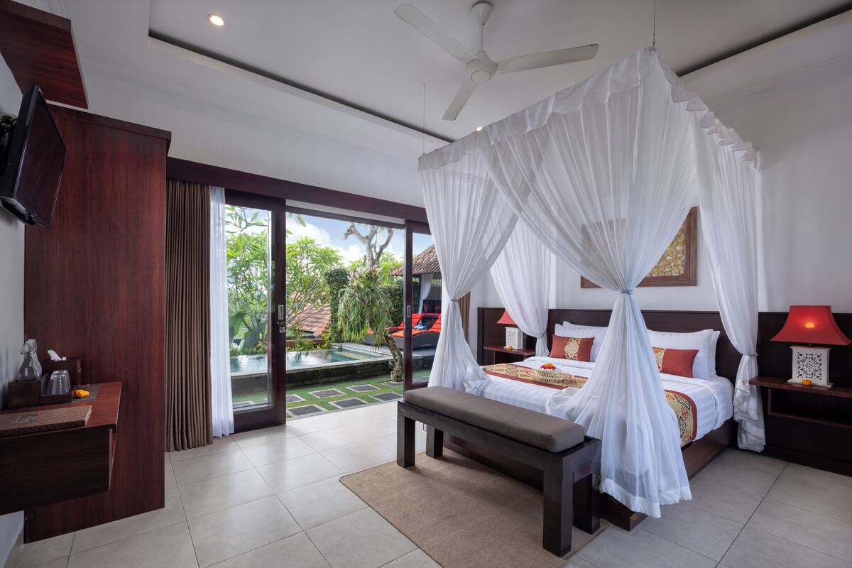 A bedroom with a bed under a canopy, a bench, a bedside table, a TV, and a view of a pool and garden.