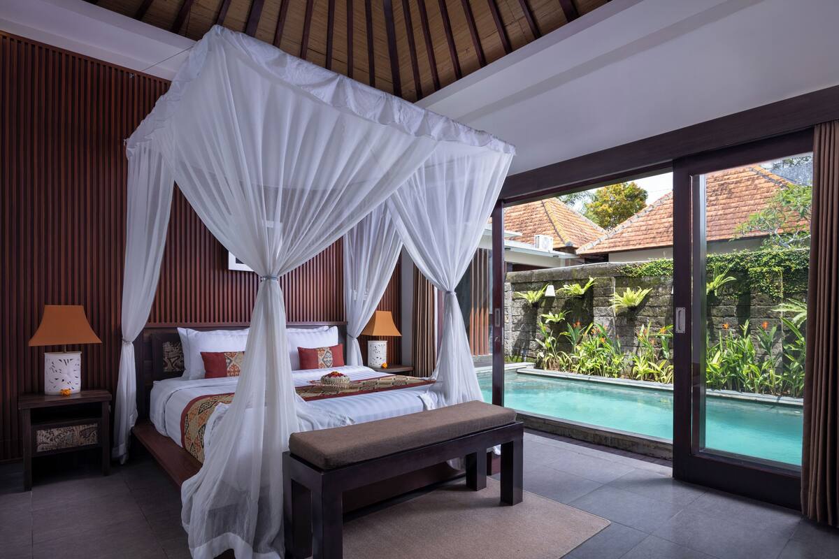 A bedroom with a four-poster bed, a bench, a nightstand with a lamp, and a view of an infinity pool.