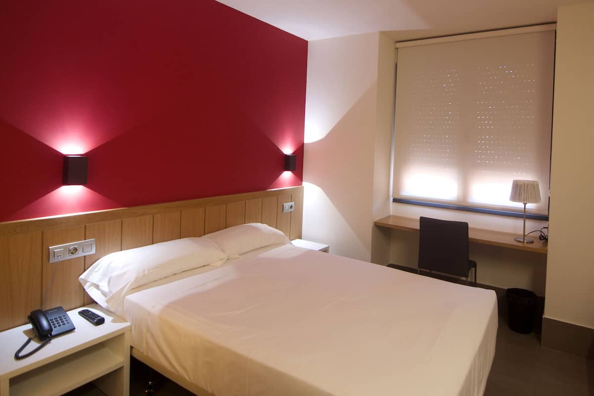 A hotel room with a bed, bedside telephone, and a desk with a chair.