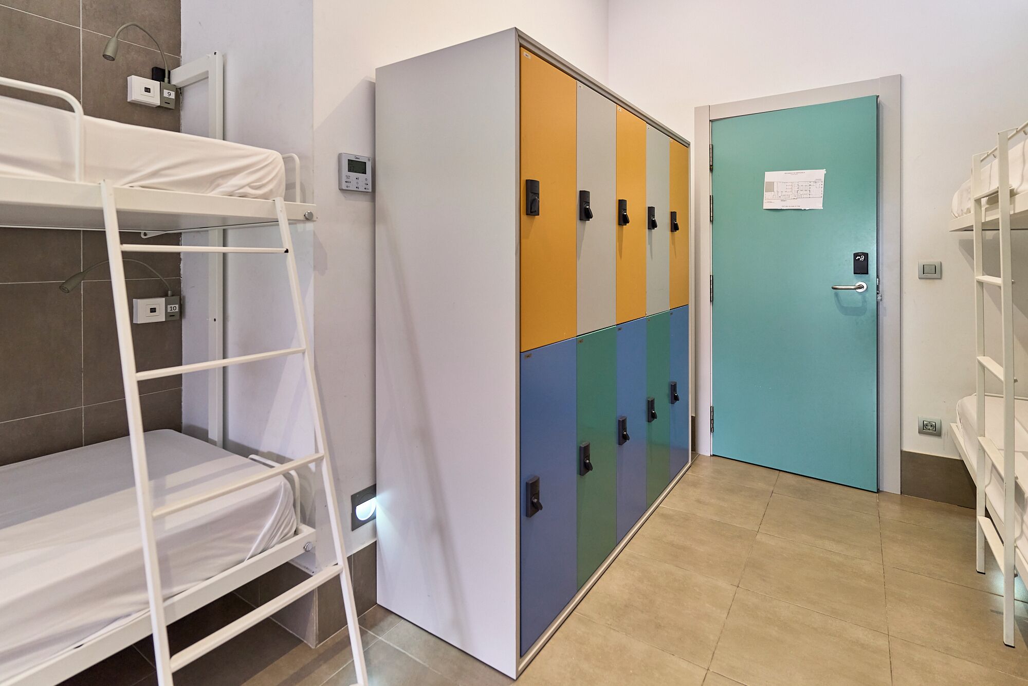 Un couloir d’hôtel avec des lits superposés, une rangée de casiers et une porte avec une pancarte.