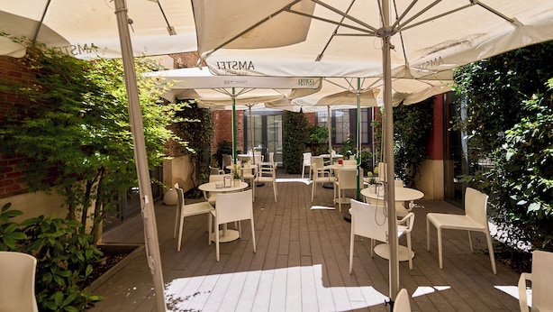 An outdoor patio area with round tables and white chairs, covered by large umbrellas, surrounded by greenery and brick walls. - The Book Factory Hostel by gaiarooms (Valladolid)