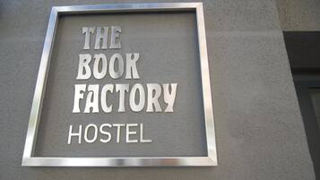 Letrero para 'The Book Factory Hostel' con estructura metálica y letras blancas sobre una pared gris.