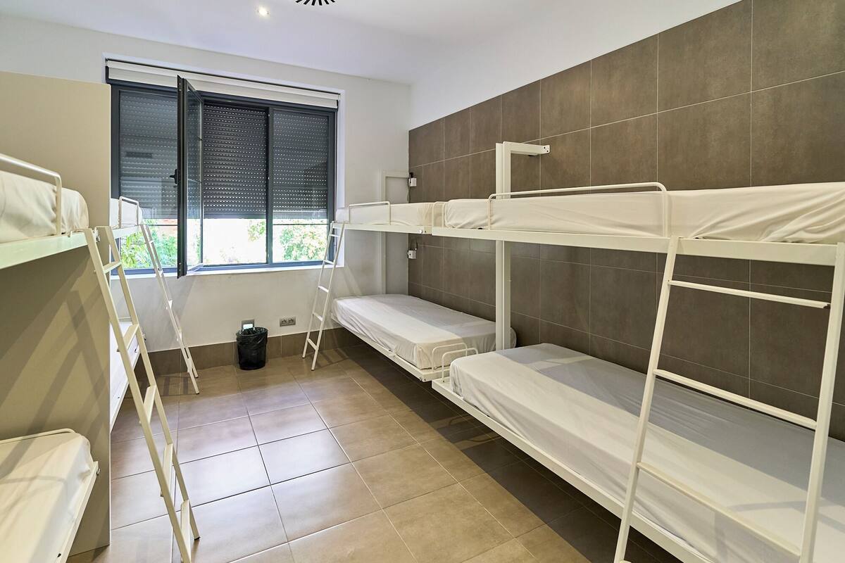 A dormitory room with bunk beds, a window with blinds, and a tiled floor.