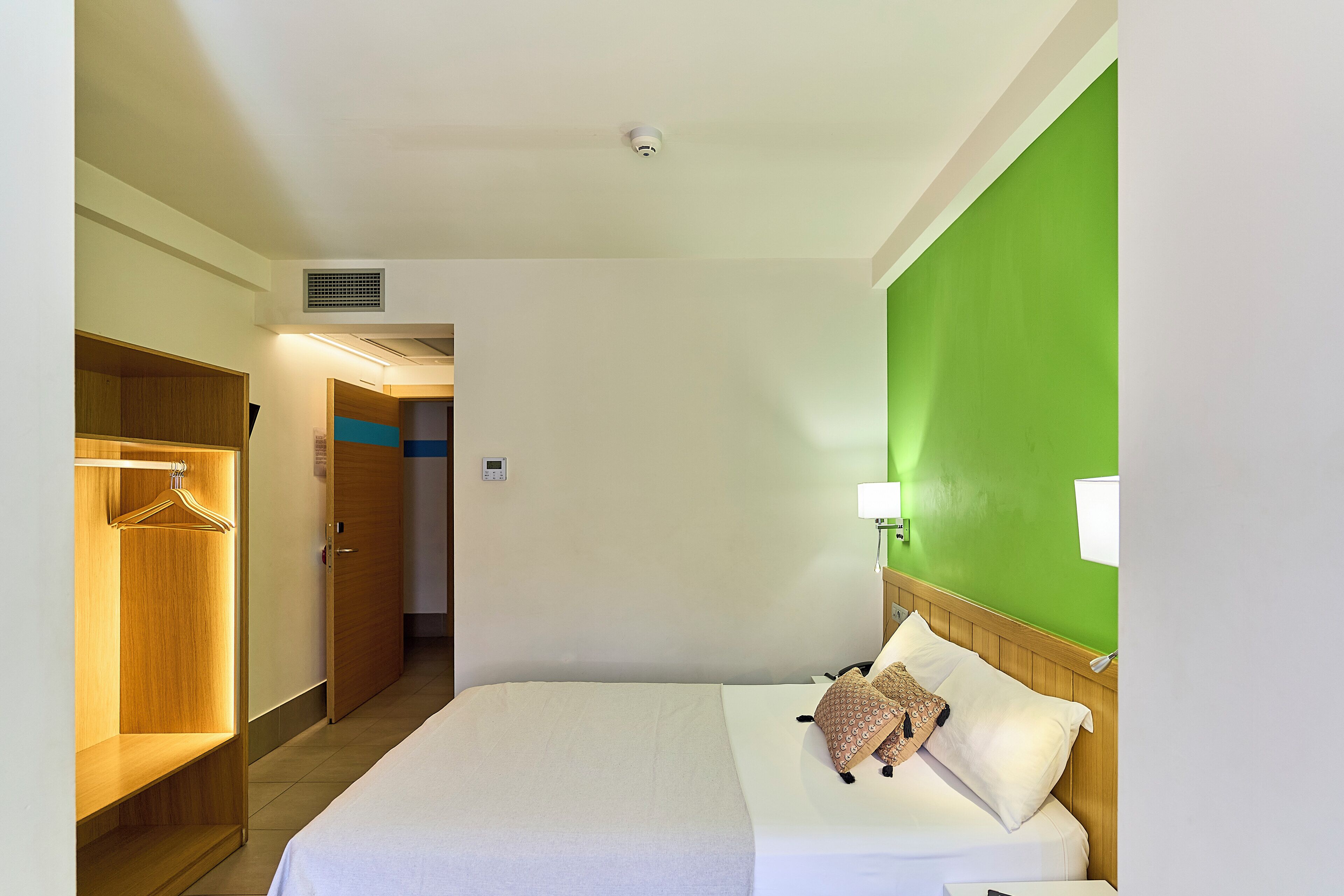 A hotel room with a bed, a wooden wardrobe, a green accent wall, and a bedside lamp.