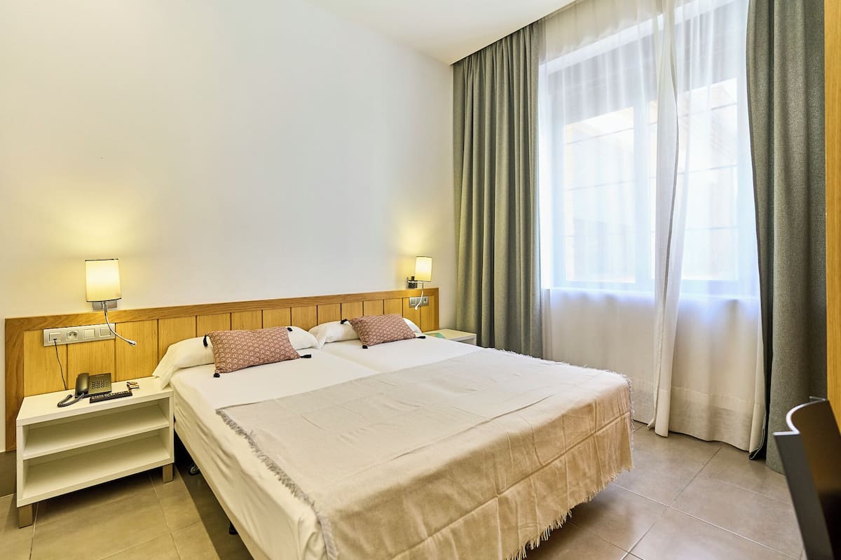 A hotel room with a large bed, two bedside tables, a nightstand, a lamp, a telephone, and a window with curtains.