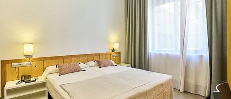 A hotel room with a large bed, two bedside tables, a nightstand, a lamp, a telephone, and a window with curtains.