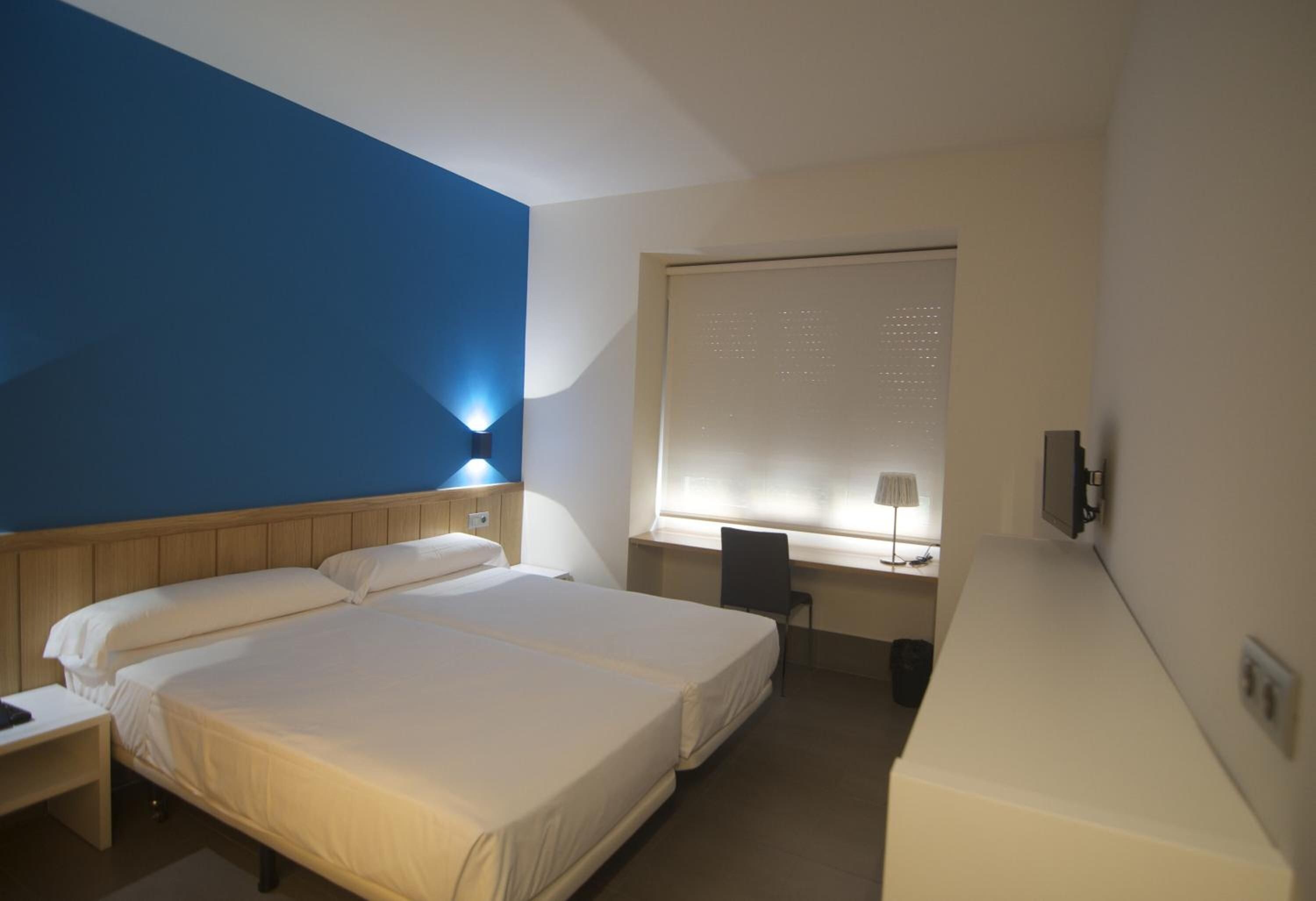 Et hotelværelse med en stor seng, et skrivebord, en stol og et vindue med persienner.