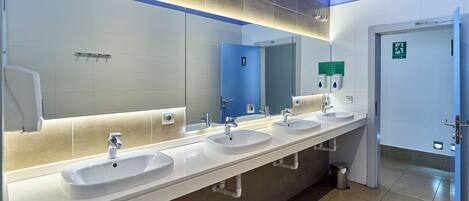 Eine öffentliche Toilette mit drei Waschbecken, einem großen Spiegel und einer blauen Tür mit einem Notausgangsschild.