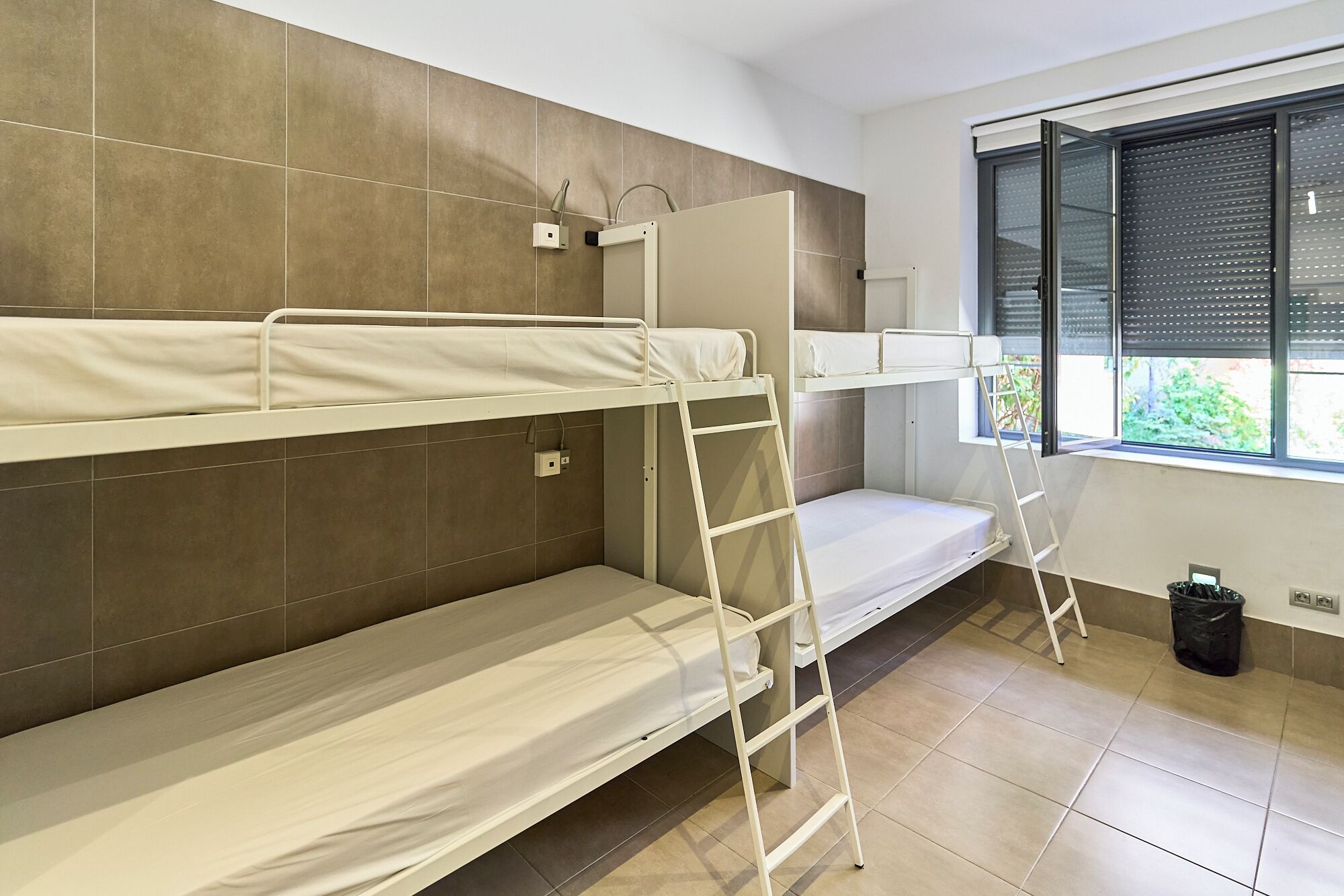 A dormitory room with bunk beds, a window with blinds, and a tiled floor.