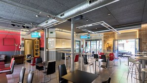 A modern restaurant with a mix of indoor and outdoor seating, exposed ductwork on the ceiling, and a red accent wall. - The Book Factory Hostel by gaiarooms (Valladolid)