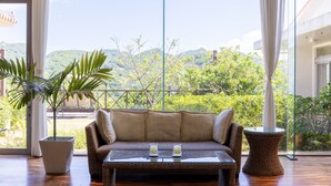 Un sofá de mimbre con una mesa de vidrio y dos vasos, ubicado en una habitación con ventanas amplias que dan a vistas de vegetación y colinas.