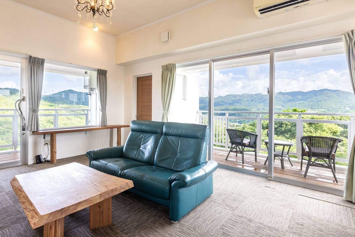 Une pièce spacieuse avec un canapé en cuir, une table en bois et un balcon offrant une vue sur des collines verdoyantes.