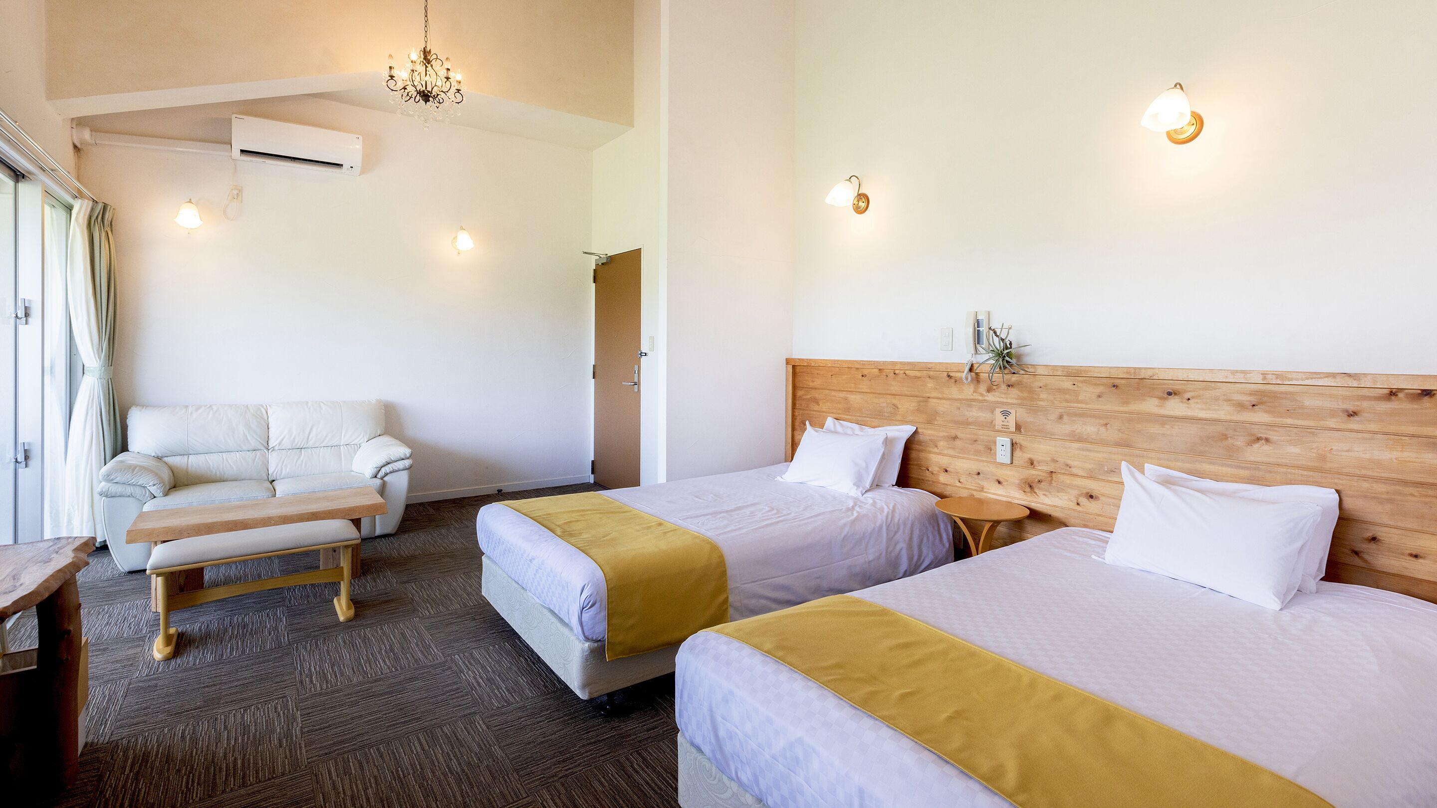 A hotel room with two beds, a sofa, a wooden headboard, a small table, and a chandelier.