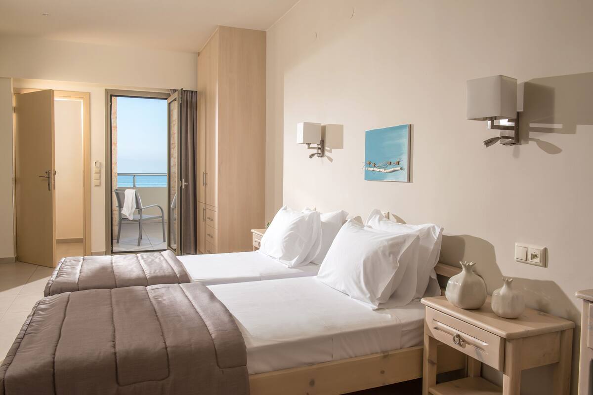 Een hotelkamer met twee bedden, een nachtkastje, een wandlamp, een schilderij en uitzicht op zee door een geopende deur.
