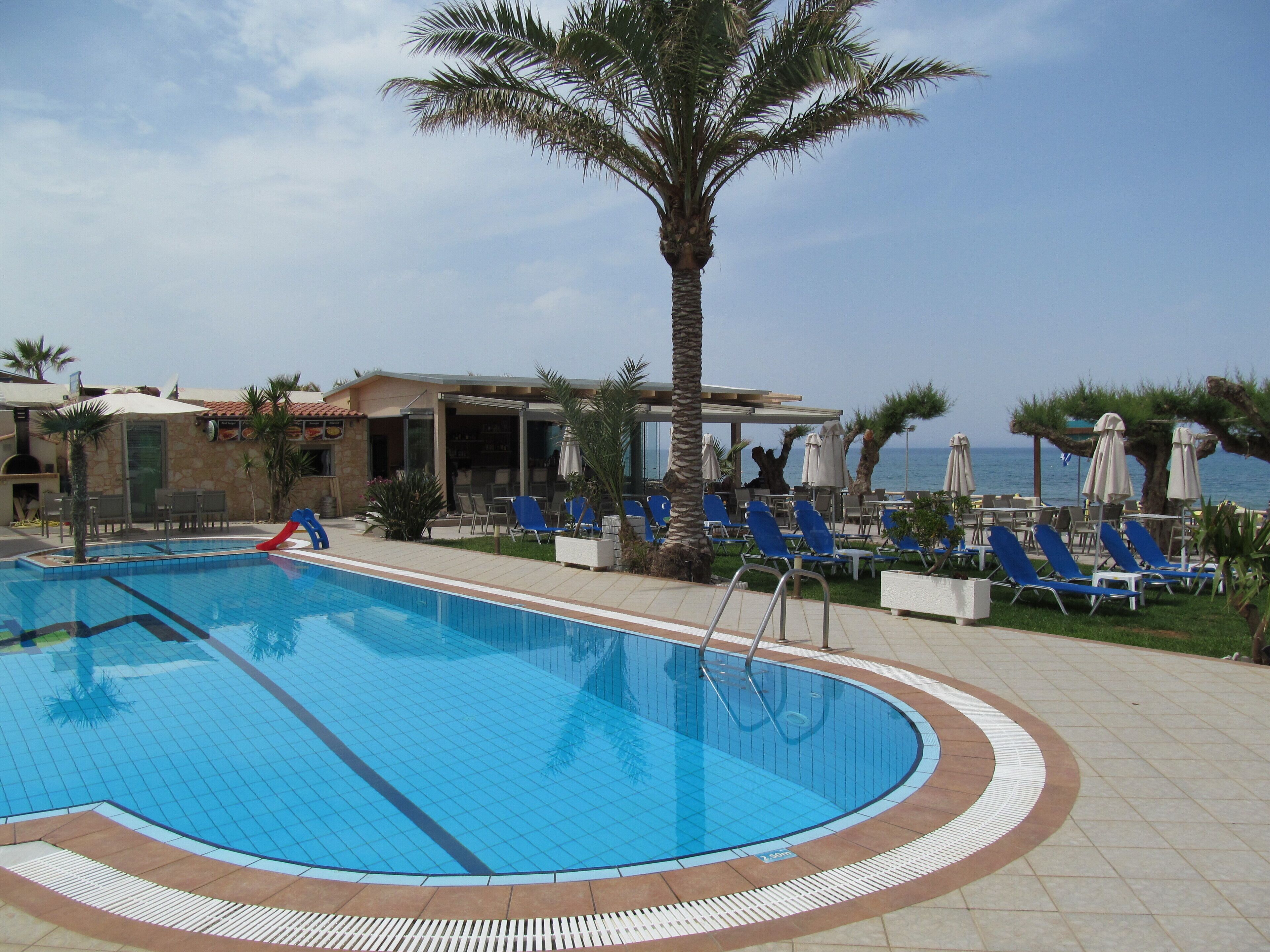 A swimming pool with blue tiles, surrounded by lounge chairs and umbrellas, with a view of the sea.