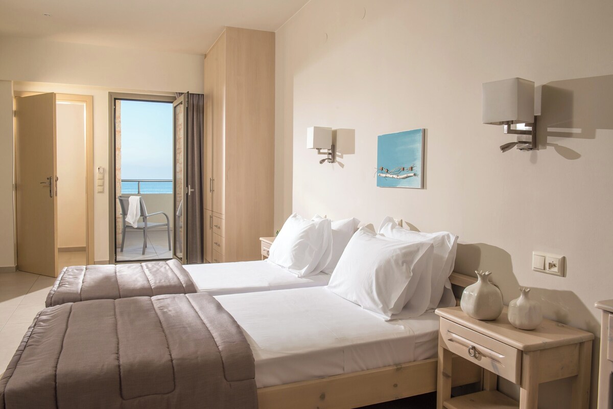 Ein Hotelzimmer mit zwei Betten, einem Nachttisch, einer Wandlampe, einem Gemälde und Meerblick durch eine offene Tür.