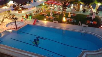 Una piscina con un logotipo, rodeada de sillones de descanso y sombrillas, y un área de comedor al fondo.