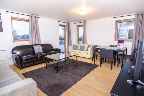 A modern living room with a black leather sofa, a glass coffee table, a beige sofa, and a dining area with a table and chairs.