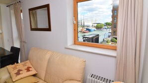 Una sala de estar con un sofá color beige, una ventana que ofrece vistas a barcos y edificios, un espejo y un radiador.