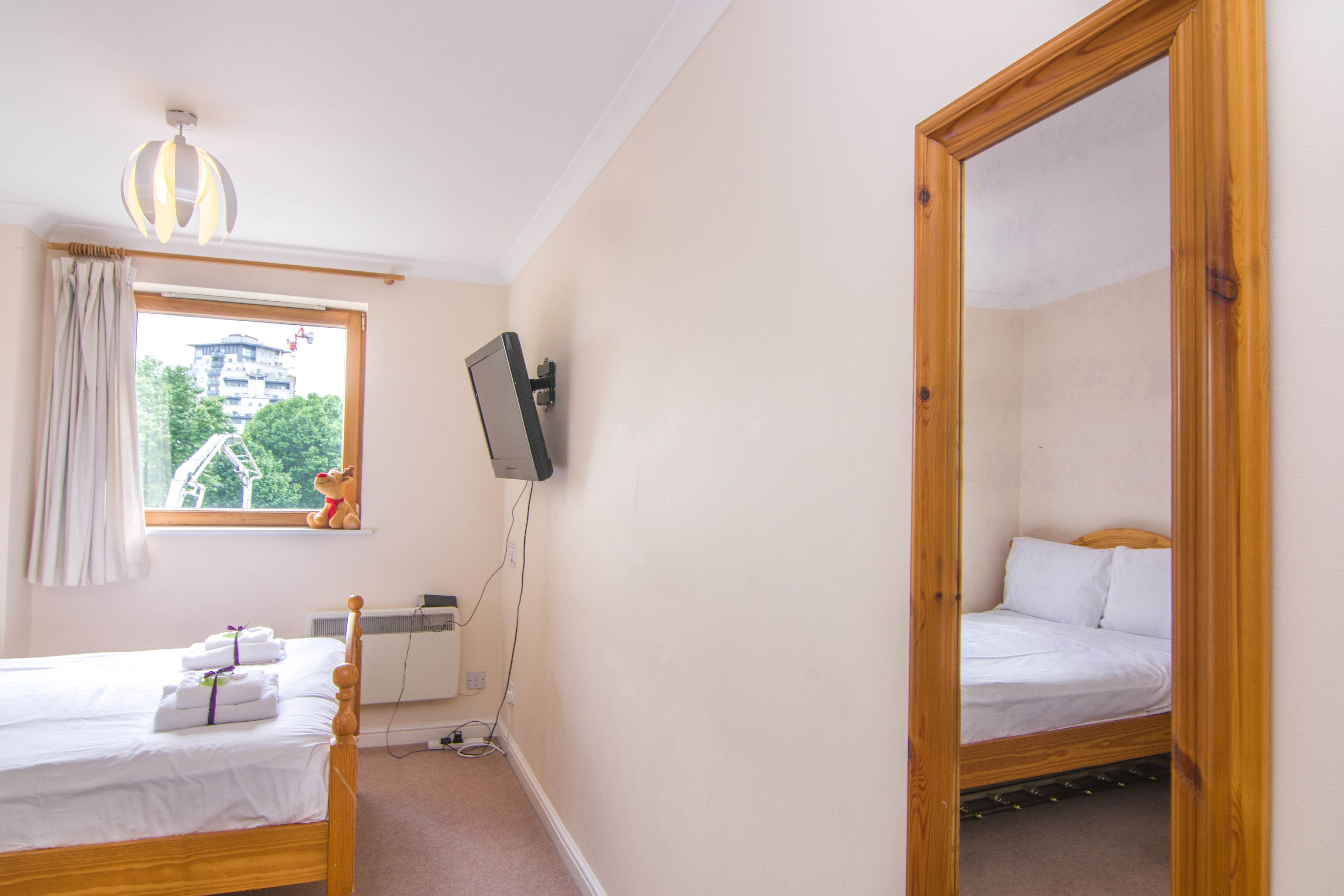 A bedroom with two beds, a TV mounted on the wall, and a window with a view of buildings and trees.