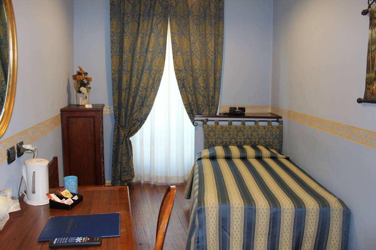 Une chambre d’hôtel comprenant un lit, un bureau avec une chaise, une petite table avec un vase de fleurs, une machine à café et une fenêtre avec des rideaux.