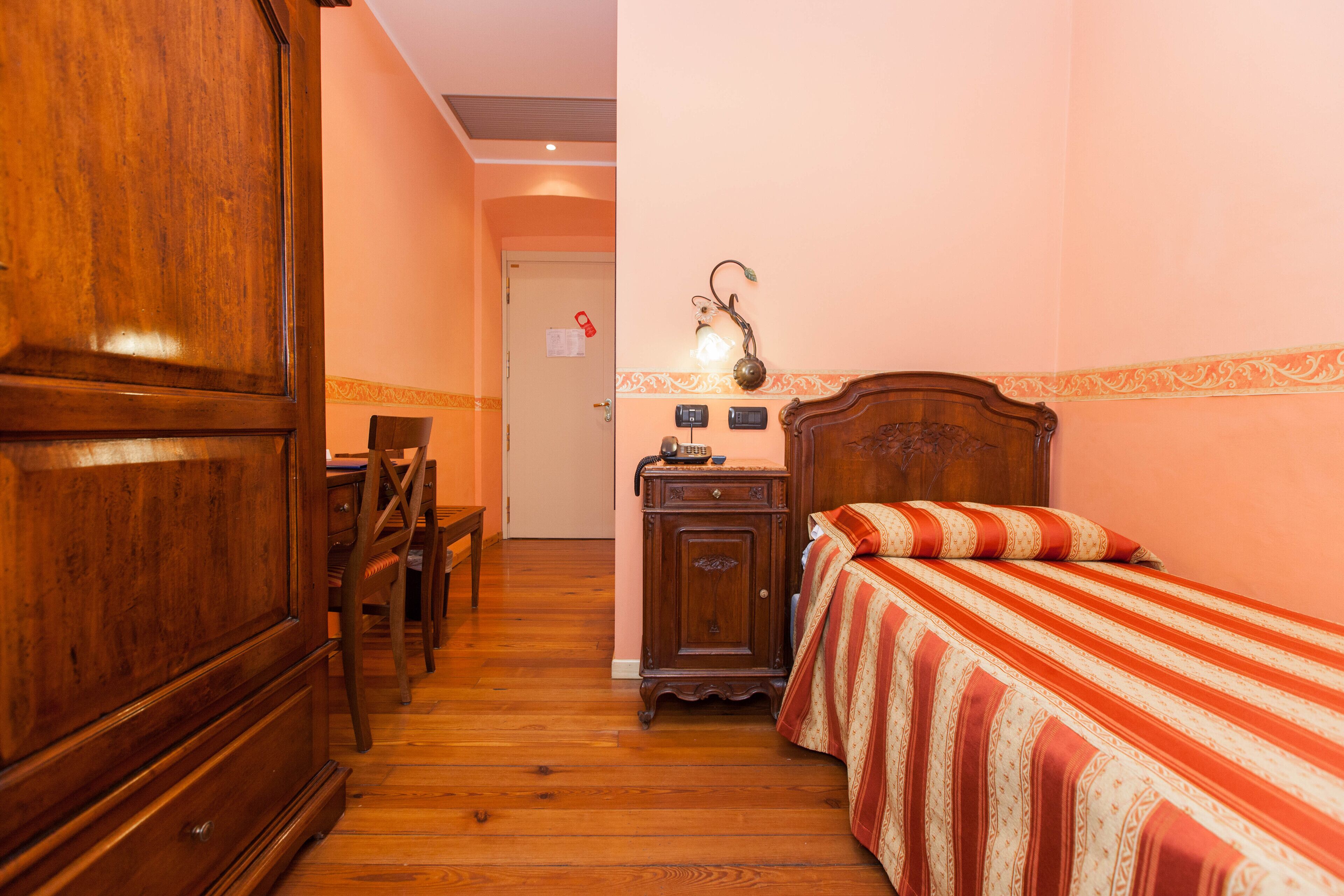 Una habitación de hotel con un armario de madera, una cama con colcha a rayas, una mesita de noche con lámpara y una silla de madera.