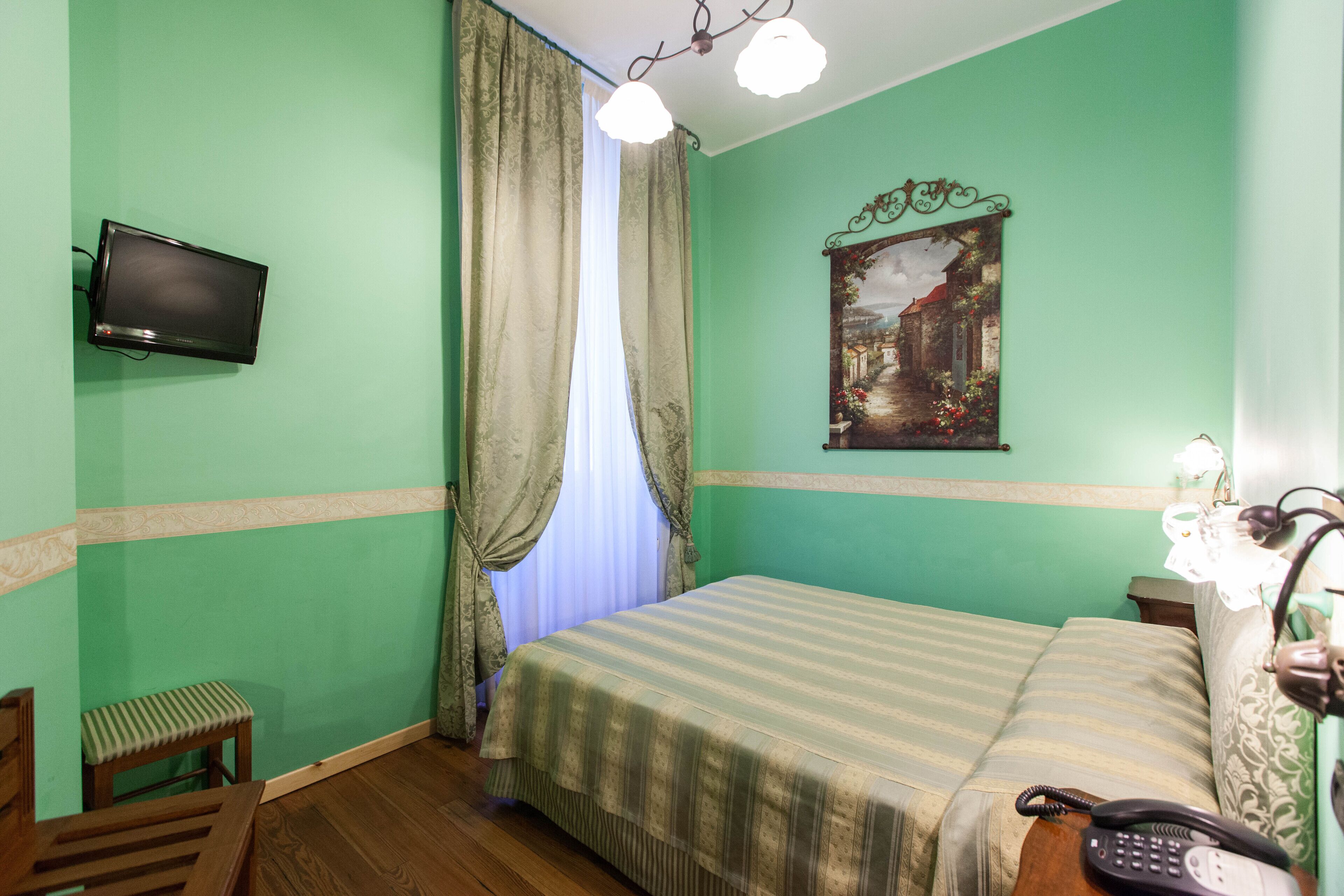 A room with a bed, a chair, a desk with a telephone, a television mounted on the wall, and a painting above the bed.