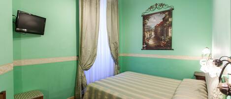 Quarto com cama, uma cadeira, escrivaninha com telefone, televisão na parede e um quadro sobre a cama.