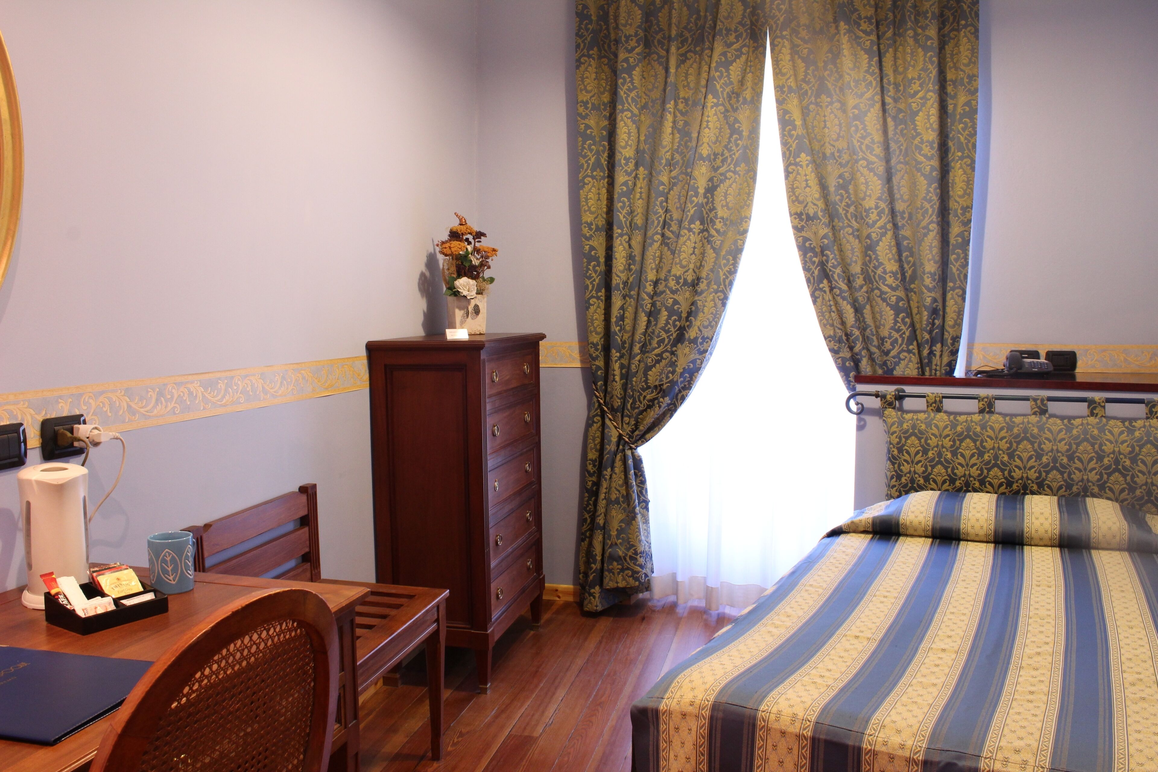 A hotel room with a wooden desk, a dresser, a bed with striped bedding, and a window with patterned curtains.