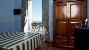 Un dormitorio con una cama, un armario de madera, un escritorio con una taza y vistas al mar a través de una ventana con cortinas.