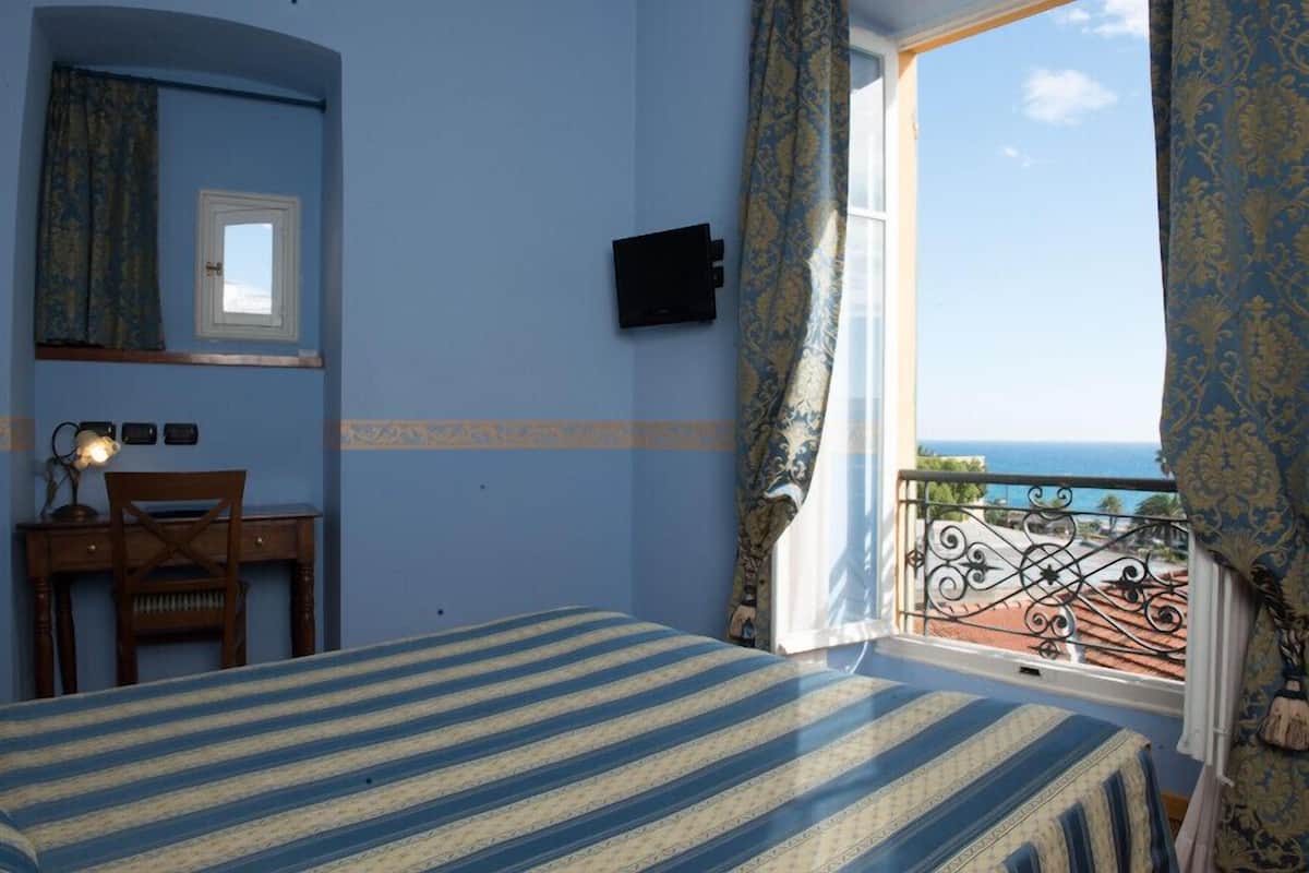Une chambre à coucher avec un lit, un bureau et une chaise. Il y a une fenêtre avec des rideaux et une vue sur la mer.
