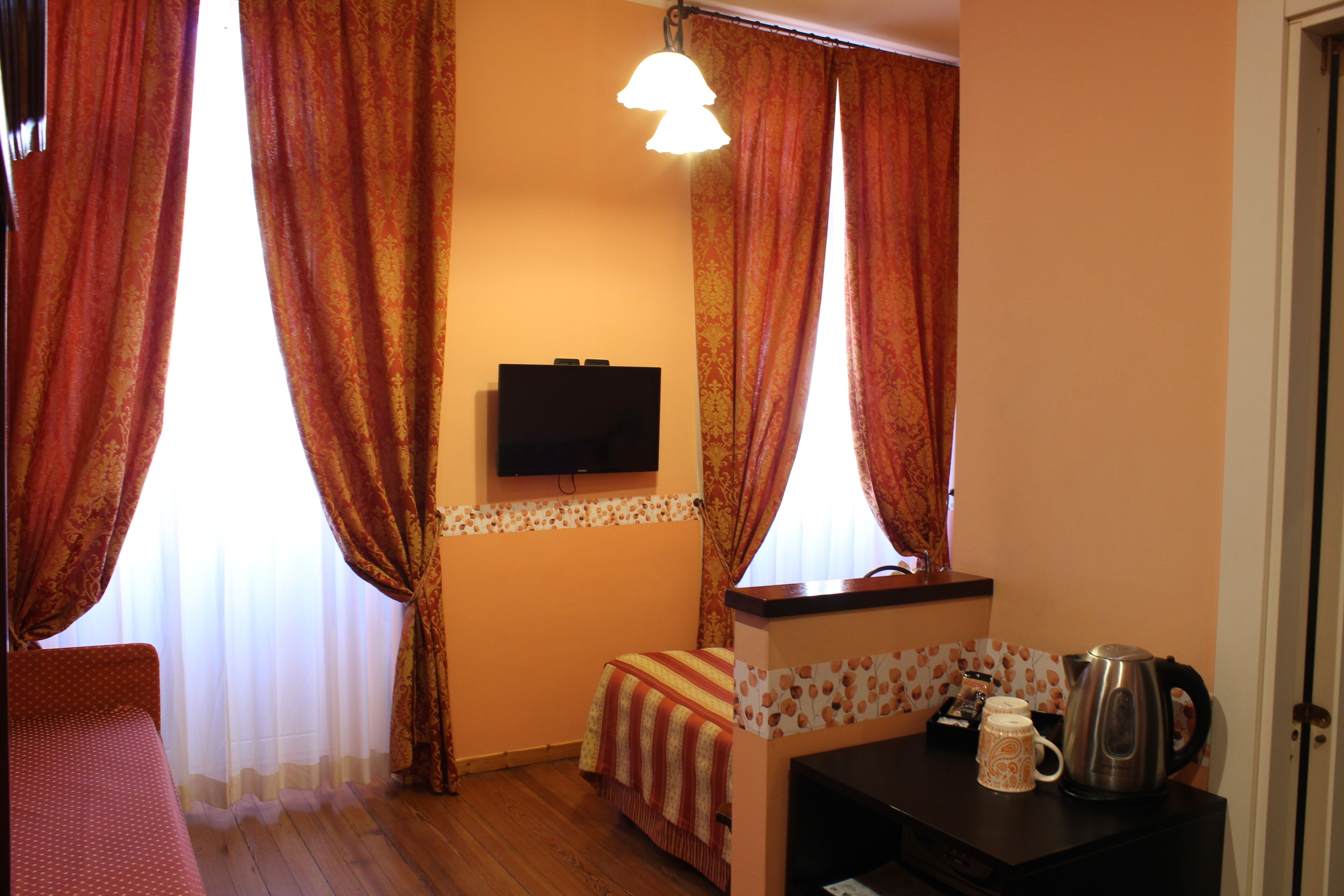 Una habitación con cama, televisor montado en la pared, una mesita con hervidor y taza, y cortinas.
